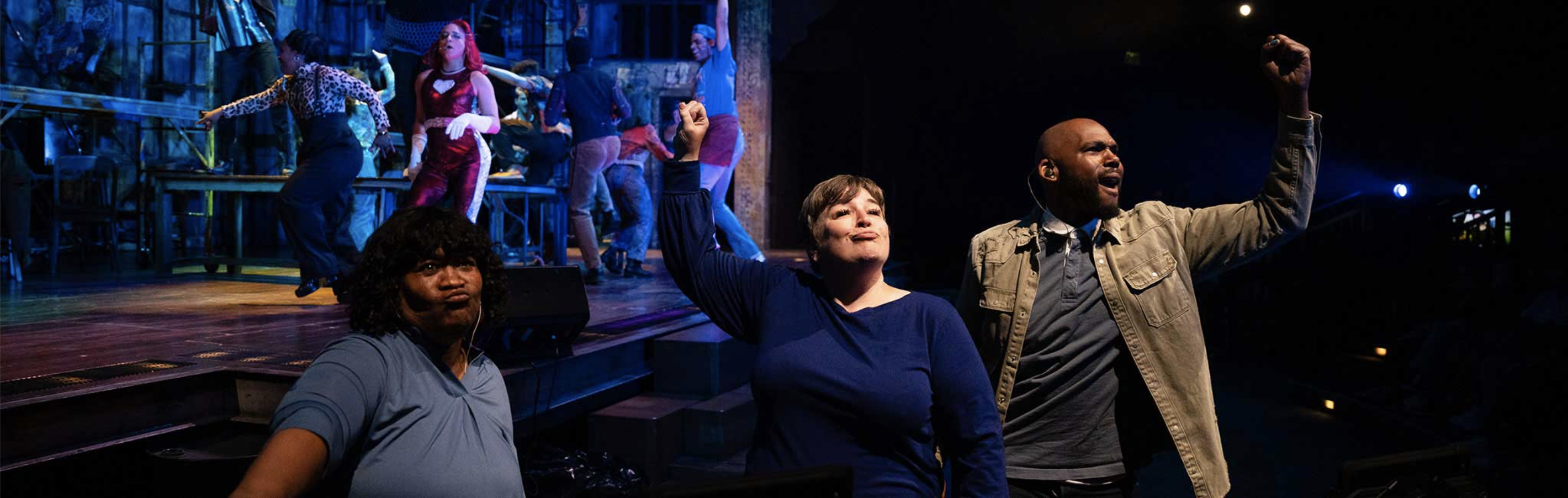 A production still from Lizard Boy by Oregon Shakespeare Festival. Three sign interpreters interpret a show front stage. Two interpreters, one white and one Black, hold their fists high in the air. A third Black interpreter stands to their left. Behind them, actors perform a choreographed musical number on stage.